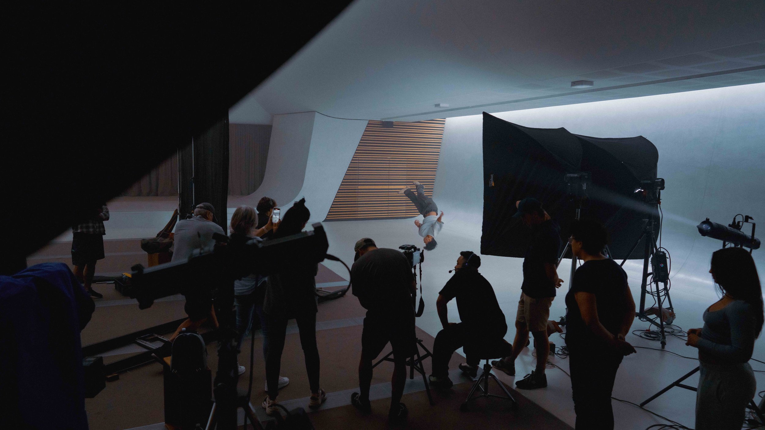 Film crew on a white studio stage, a performer inverted above the floor, cameras in the foreground.